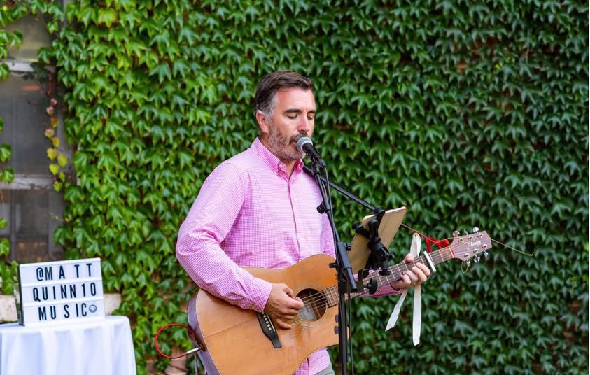 Matthew in a pink shirt singing and playing acoustic guitar against a leafy backdrop