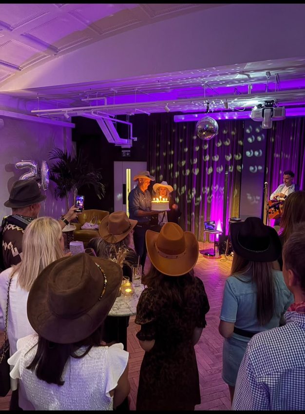 Guests in cowboy hats gathered around a birthday cake, with Matthew performing guitar to the side