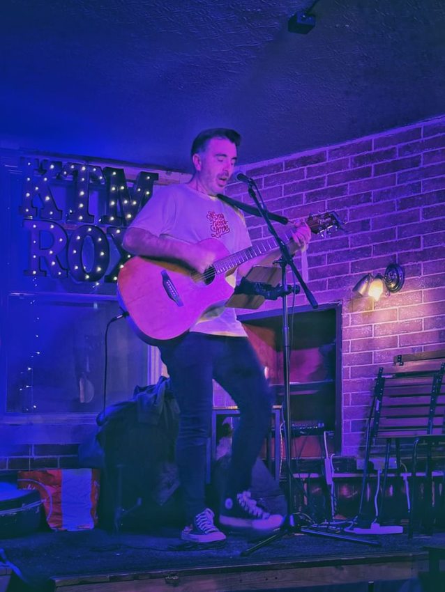 Matthew singing and playing acoustic guitar on a small stage under blue and purple lights