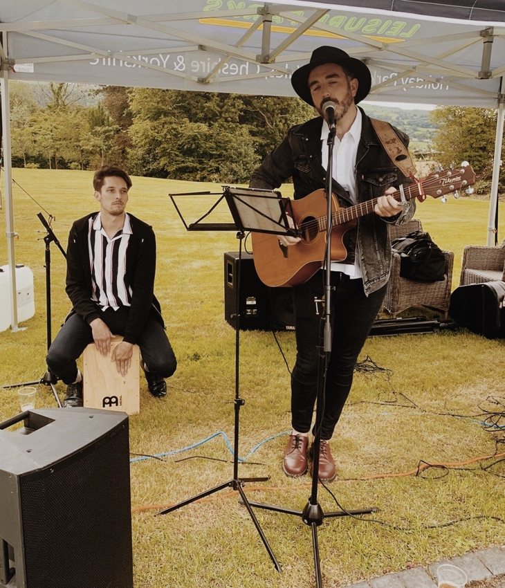 Matthew singing and playing guitar with a cajon percussionist under an outdoor canopy