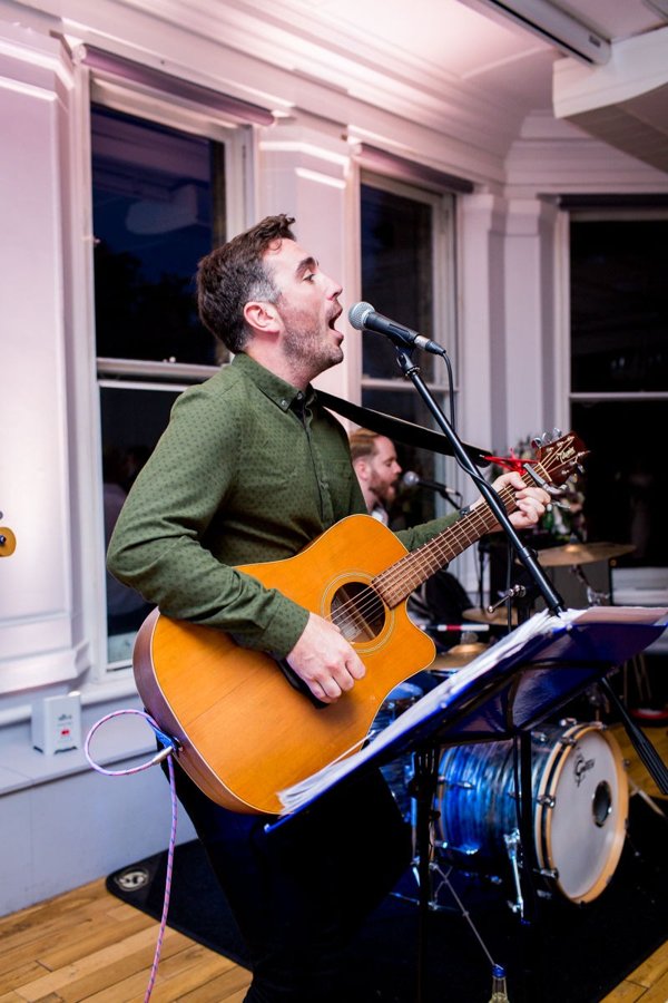 Matthew singing and playing acoustic guitar with a drummer performing behind him