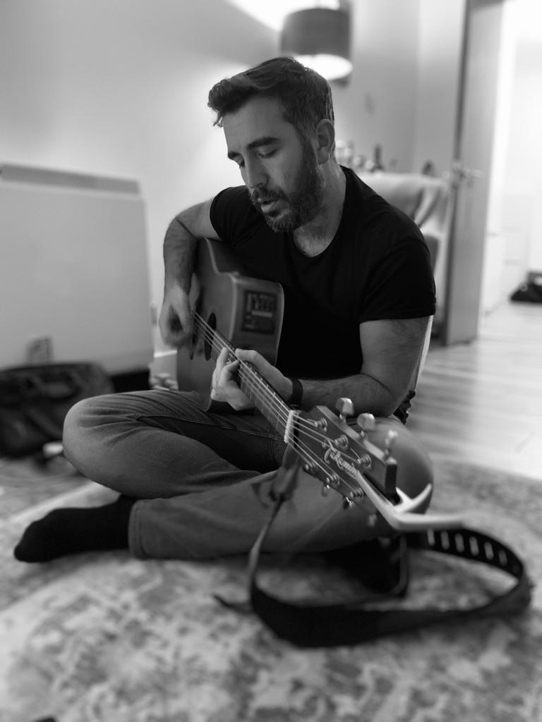 Black-and-white rehearsal photograph of Matthew seated on the floor playing acoustic guitar